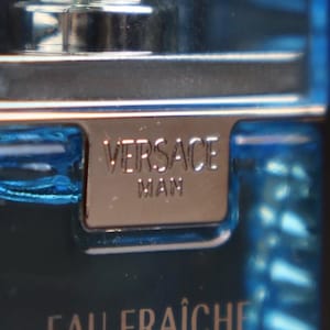 Puede incluir: Primer plano de una botella de vidrio azul de colonia Versace Man Eau Fra&icirc;che. La etiqueta de metal plateado dice "VERSACE MAN". La botella tiene una tapa plateada y las palabras "EAU FRA&Icirc;CHE" son visibles.