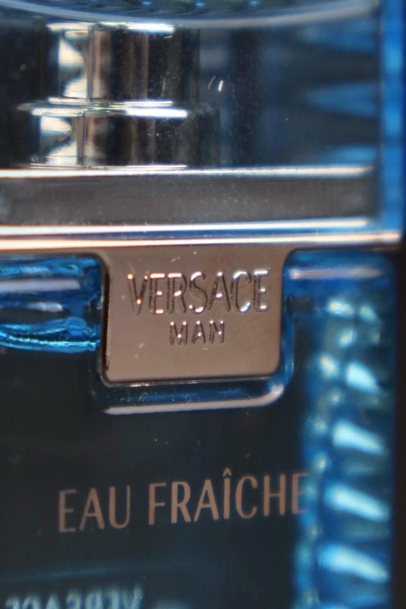 Puede incluir: Primer plano de una botella de vidrio azul de colonia Versace Man Eau Fra&icirc;che. La etiqueta de metal plateado dice "VERSACE MAN". La botella tiene una tapa plateada y las palabras "EAU FRA&Icirc;CHE" son visibles.