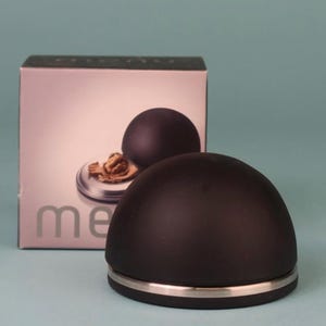 May include: A black nutcracker with a silver band at the base, next to a box with a similar nutcracker and a walnut. The word "me" is visible on the box. The nutcracker has a rounded, dome-like shape.