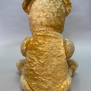 May include: A vintage, golden-brown teddy bear with a plush, fuzzy texture. The bear is sitting with its back to the camera, showing its stitched seams.