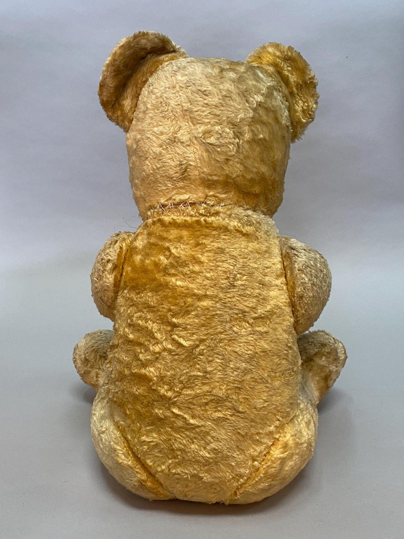 May include: A vintage, golden-brown teddy bear with a plush, fuzzy texture. The bear is sitting with its back to the camera, showing its stitched seams.