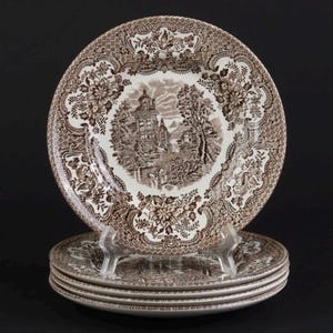 Może przedstawiać: Stos czterech okrągłych talerzy ceramicznych z brązowo-białym wzorem transferowym. Środek każdego talerza przedstawia szczegółowy krajobraz z budynkiem i drzewami, otoczony szeroką bordiurą z motywami kwiatowymi oraz geometrycznymi wzorami.