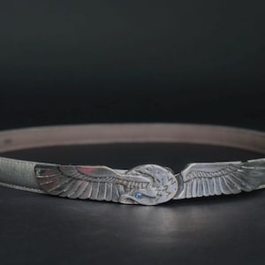 Op de afbeelding: Zilverkleurige riem met een adelaarvormige gesp. De gesp heeft een adelaar met uitgespreide vleugels en een kleine blauwe edelsteen. De riem is lichtgrijs en lijkt van leer te zijn.