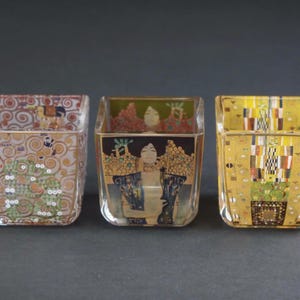 Op de afbeelding: Drie vierkante glazen kaarsenhouders met kleurrijke artistieke prints. De linkerhouder toont wervelende gouden en groene patronen. De middelste houder toont een portret van een persoon met donker haar. De rechterhouder heeft een geometrisch goudontwerp.