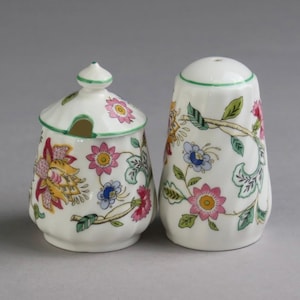 May include: A white porcelain sugar bowl with a lid and a matching salt shaker, both featuring a colourful floral pattern with pink, yellow, and blue flowers, green leaves, and thin green trim along the edges of the lid and the top rim.
