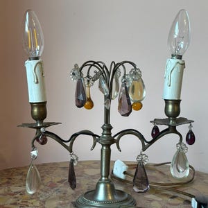May include: An antique candelabra with two faux candle lights. The metal fixture has a bronze finish and is adorned with clear, purple, and yellow crystal droplets. The base is round and the bulbs are clear.