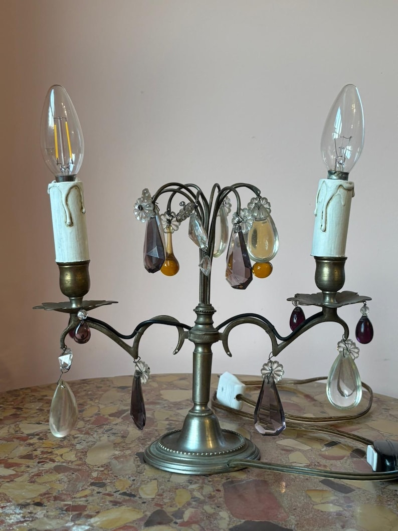 May include: An antique candelabra with two faux candle lights. The metal fixture has a bronze finish and is adorned with clear, purple, and yellow crystal droplets. The base is round and the bulbs are clear.