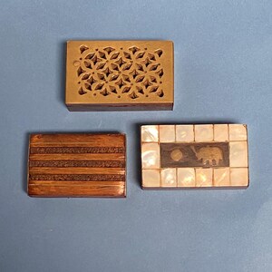 Könnte beinhalten: Drei kleine Holzkisten mit aufwendigen Designs. Die erste Kiste hat ein geometrisches Muster, das in das Holz geschnitzt ist. Die zweite Kiste hat ein gestreiftes Muster. Die dritte Kiste hat eine Perlmutteinlage mit einem Elefantenmotiv.