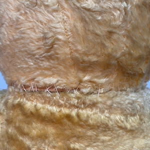 May include: Close-up of the back seam of a light brown plush toy. The seam is stitched with a light brown thread.