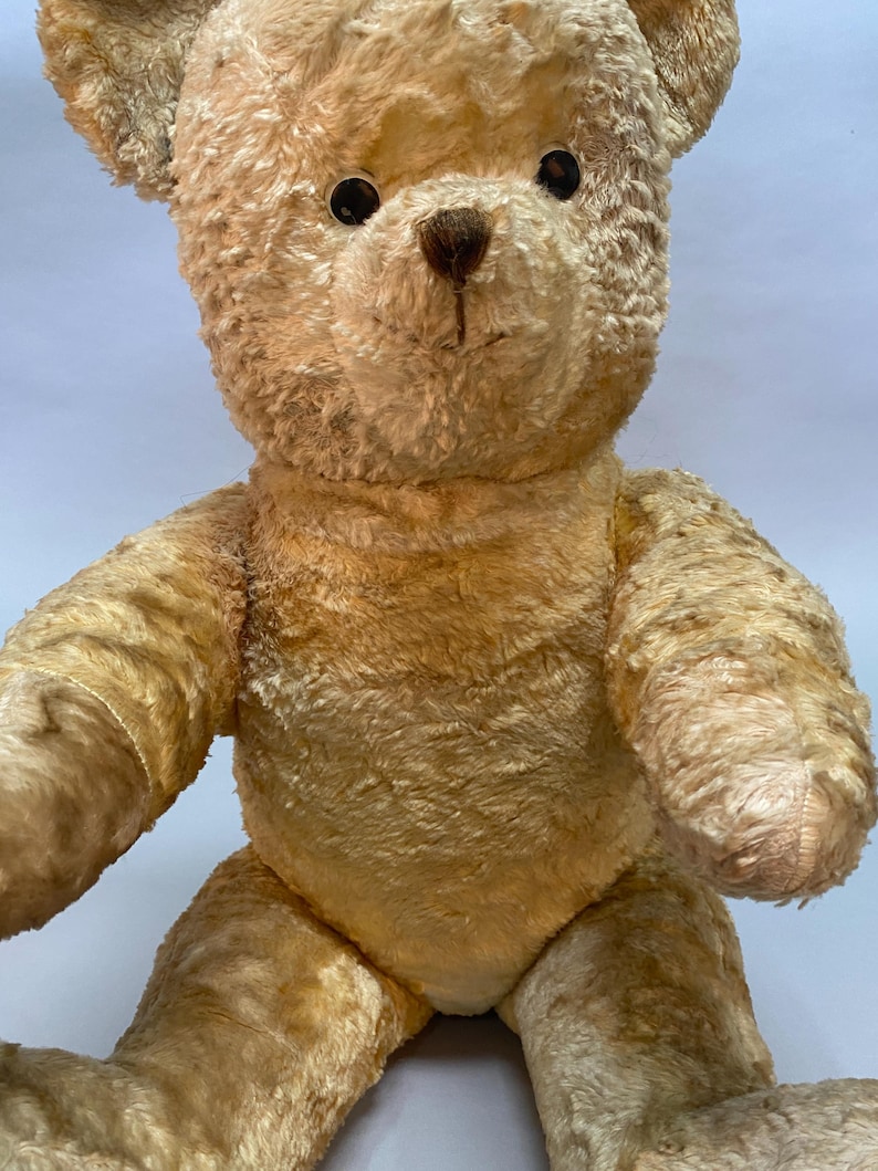 May include: A vintage, tan teddy bear with black button eyes and a brown nose. The bear is sitting with its arms outstretched.