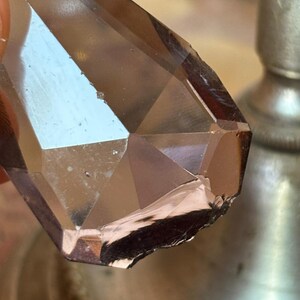 May include: Close-up of a faceted, brown crystal with a geometric design. The crystal has a polished surface with multiple reflective planes. The crystal is partially chipped on one edge, revealing a rougher texture. The background is blurred.