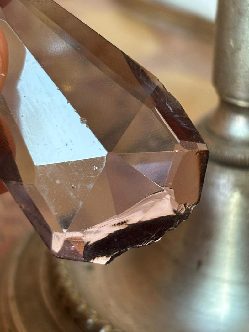 May include: Close-up of a faceted, brown crystal with a geometric design. The crystal has a polished surface with multiple reflective planes. The crystal is partially chipped on one edge, revealing a rougher texture. The background is blurred.