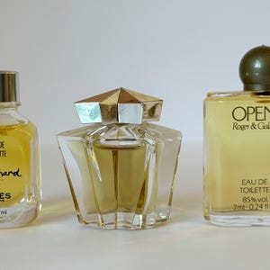 以下が含まれることがあります： 3つのヴィンテージ香水瓶。左側のボトルには「Cabochard」と「Eau de Toilette」と書かれています。中央のボトルは、金と銀のキャップが付いた幾何学的なデザインです。右側のボトルには「Open」と「Eau de Toilette」と書かれており、濃い緑色のキャップが付いています。ボトルには黄色の液体が入っています。