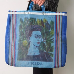 Op de afbeelding: Blauwe mesh tote bag met een Frida Kahlo portret en groene bladeren. De tas heeft blauwe handvatten en een witte rand. Het woord "FRIDA" staat onderaan gedrukt.