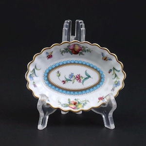 Pode incluir: Um prato oval de porcelana com borda festonada e acabamento dourado. O centro apresenta um motivo floral rodeado por uma borda festonada azul-clara. A superfície branca é decorada com frutas coloridas, pequenas flores e ilustrações de borboletas.