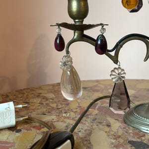 May include: A vintage brass candelabra with a white candle, adorned with hanging crystal and ruby-colored teardrop accents. The lamp features a black and white on/off switch and a white electrical plug.