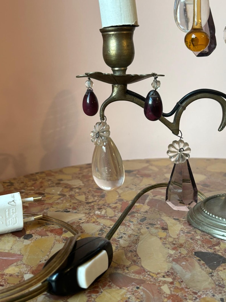 May include: A vintage brass candelabra with a white candle, adorned with hanging crystal and ruby-colored teardrop accents. The lamp features a black and white on/off switch and a white electrical plug.
