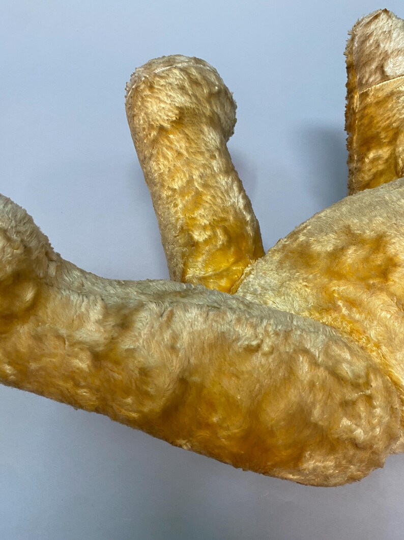 May include: A vintage, yellow plush teddy bear with a worn, faded appearance. The bear is lying on its back with its legs in the air.