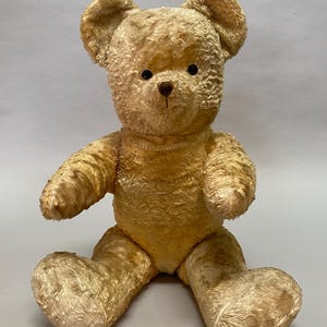 May include: A vintage, plush, beige teddy bear with black eyes and a brown nose. The bear is sitting with its arms outstretched and its legs bent.