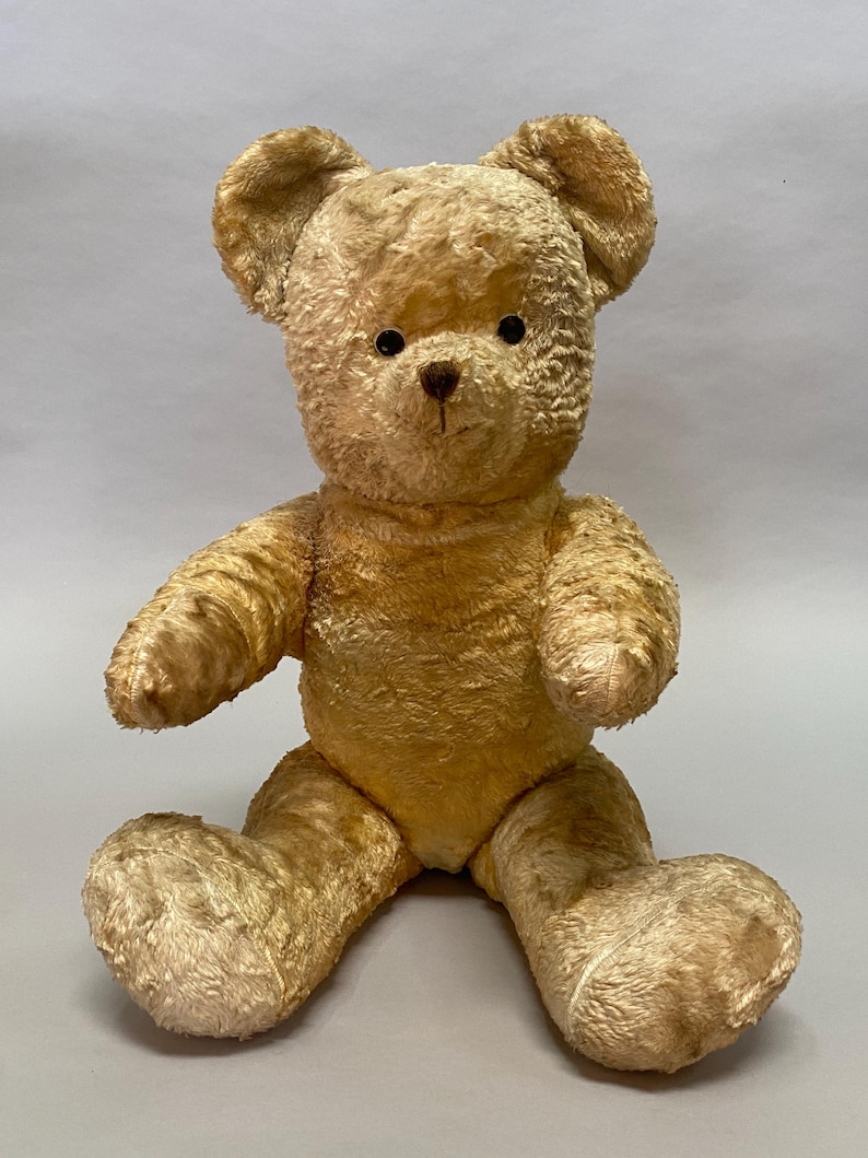 May include: A vintage, plush, beige teddy bear with black eyes and a brown nose. The bear is sitting with its arms outstretched and its legs bent.