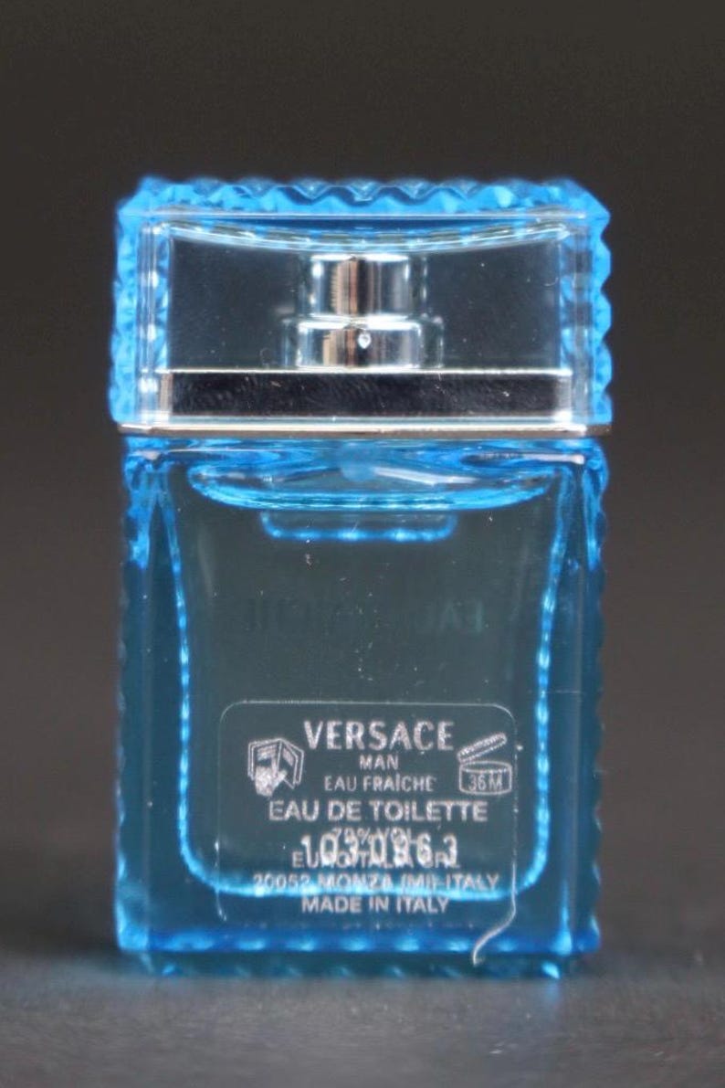 Puede incluir: Peque&ntilde;a botella rectangular de Versace Man Eau Fraiche Eau de Toilette. La botella de vidrio azul transparente tiene un atomizador plateado y una tapa azul claro texturizada. La etiqueta incluye el nombre de la marca y detalles del producto.