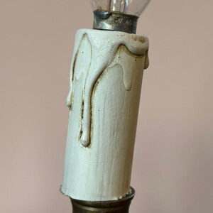 May include: Close-up of a vintage-style light fixture. The fixture features a cream-colored, candle-shaped base with dripping wax details. A clear glass light bulb is screwed into the top, and the base sits on a brass-colored metal fixture.