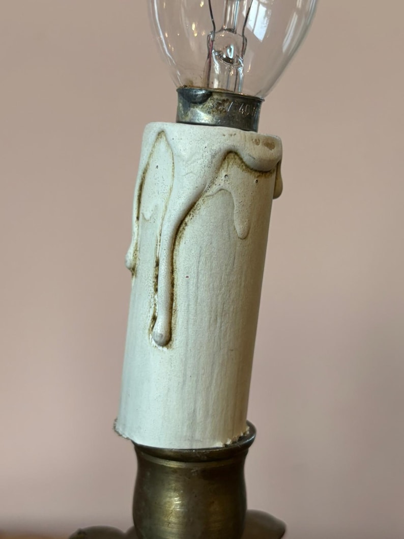 May include: Close-up of a vintage-style light fixture. The fixture features a cream-colored, candle-shaped base with dripping wax details. A clear glass light bulb is screwed into the top, and the base sits on a brass-colored metal fixture.