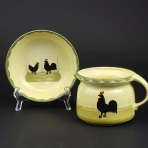 May include: A cream-coloured ceramic bowl and a matching jug, both featuring a hand-painted black rooster motif with red combs. The bowl has a scalloped green rim, while the jug includes a side handle and a green scalloped edge.