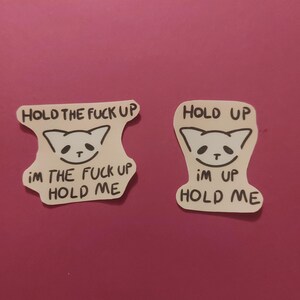 Könnte beinhalten: Zwei weiße Aufkleber mit schwarzem Text und einem Katzen-Gesicht-Design. Die Aufkleber lesen "HOLD THE FUCK UP", "i'm the fuck up hold me" und "HOLD UP", "i'm up hold me". Die Aufkleber befinden sich auf einem rosa Hintergrund.