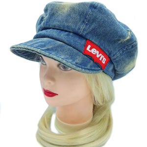 May include: A blue denim cap with a short brim and a rounded crown. The cap features a red Levi's logo patch on the side. The denim has a faded, worn look. The cap is displayed on a mannequin head with blonde hair and red lipstick.