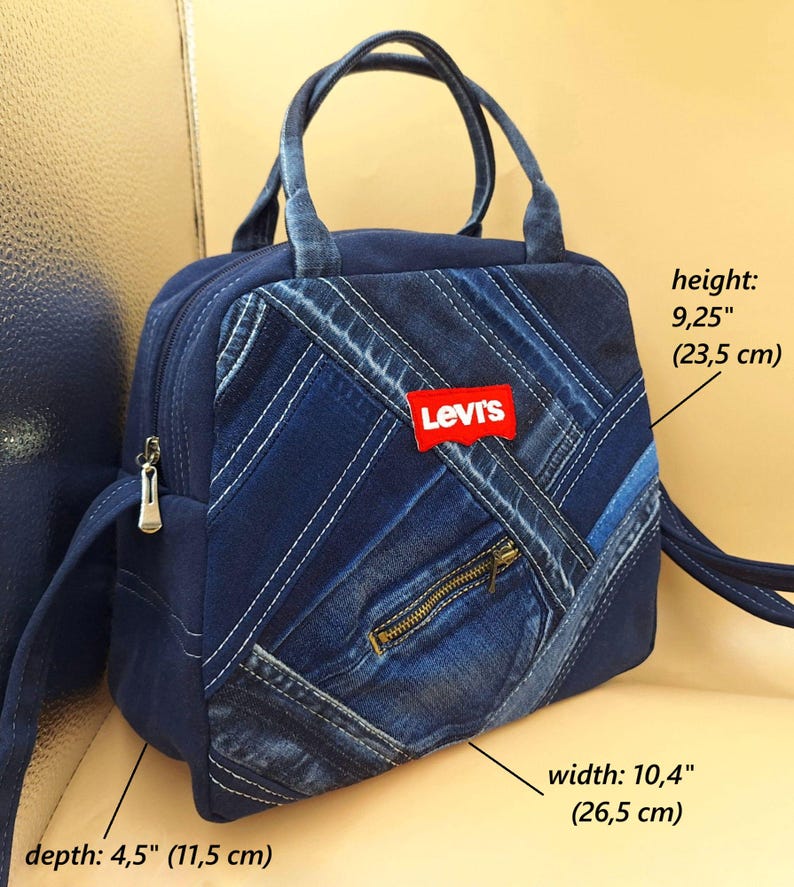 May include: A blue denim handbag featuring a patchwork design with diagonal strips, a red Levi's logo patch, and a front zipper pocket. The bag has two top handles and measures 9.25 inches in height, 10.4 inches in width, and 4.5 inches in depth.