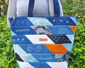Könnte beinhalten: Eine Tragetasche aus diagonalen Patchwork-Streifen aus blauem Denim, weißem Stoff und orangefarbenen Akzenten. Die Tasche verfügt über weiße Stickereien wie Sterne, Linien und Schnörkel, einen Reißverschluss oben und ein kleines rechteckiges Lederetikett.