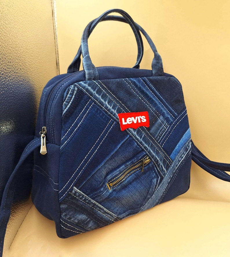 May include: A blue denim handbag featuring a patchwork design of various denim shades and textures. The bag includes two top handles, a zippered front pocket, and a red rectangular patch with the white text Levi's centered on the front panel.
