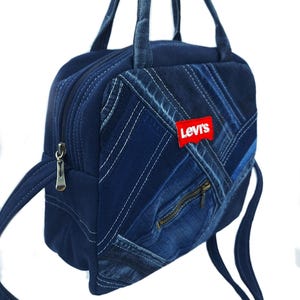 May include: A dark blue denim handbag featuring a patchwork design with visible stitching, a zippered front pocket, and a red Levi's brand patch. The bag includes two top handles and a long adjustable shoulder strap with silver metal hardware.