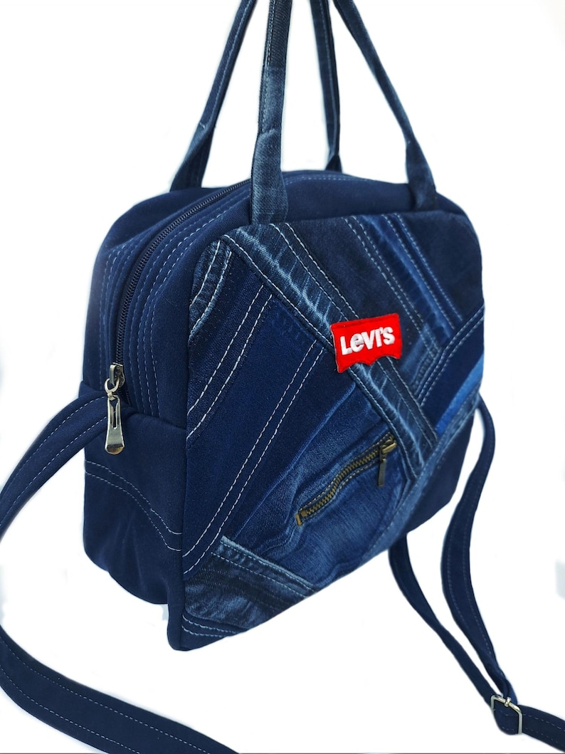 May include: A dark blue denim handbag featuring a patchwork design with visible stitching, a zippered front pocket, and a red Levi's brand patch. The bag includes two top handles and a long adjustable shoulder strap with silver metal hardware.
