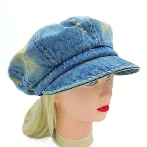 May include: A blue denim newsboy cap with a short brim. The hat has a distressed, faded appearance with light yellow patches. The cap is worn on a mannequin head with blonde hair and red lipstick.