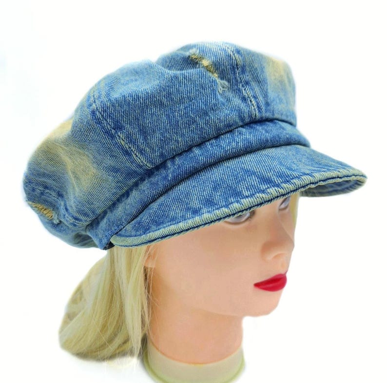 May include: A blue denim newsboy cap with a short brim. The hat has a distressed, faded appearance with light yellow patches. The cap is worn on a mannequin head with blonde hair and red lipstick.