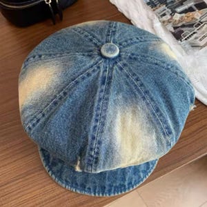 May include: A blue denim newsboy cap with a faded, distressed appearance. The cap features a rounded crown with a button detail at the top and a short, curved brim. The denim has a light blue wash with areas of lighter fading.