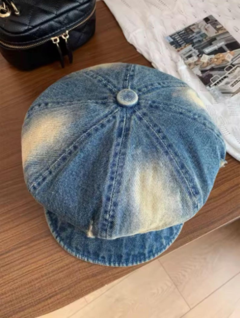 May include: A blue denim newsboy cap with a faded, distressed appearance. The cap features a rounded crown with a button detail at the top and a short, curved brim. The denim has a light blue wash with areas of lighter fading.