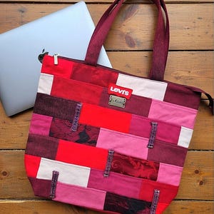May include: A patchwork tote bag constructed from various rectangular fabric pieces in shades of red, pink, and burgundy. The bag features a red Levi's brand label and a metal Levi's Jeans plaque. It has two dark burgundy shoulder straps and a zipper.