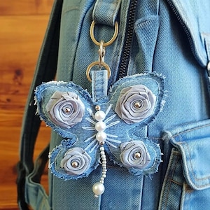 May include: A butterfly-shaped keychain crafted from blue denim fabric with frayed edges. The wings are decorated with four light blue fabric roses and a central body made of white pearls and silver beads, attached to a metal ring on a denim backpack.