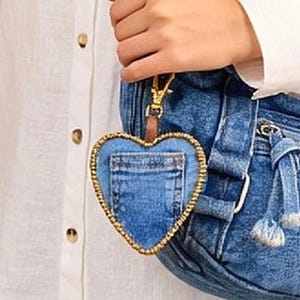 May include: A heart-shaped denim keychain featuring a small pocket on the front and a gold-toned beaded border. The charm is attached to a gold-colored metal lobster clasp and is suspended from a blue denim bag strap.