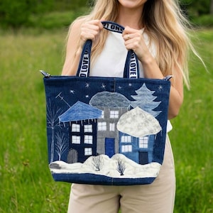 May include: A dark blue denim tote bag featuring a quilted winter village scene with houses, snow-covered ground, and a pine tree. The bag has two handles with the text "LOVE" printed in white. The design includes white embroidered stars and trees.