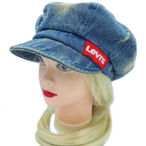 May include: A blue denim cap with a short brim. The cap has a red Levi's logo patch on the side. The denim has a faded, worn look. The cap is on a mannequin head with blonde hair and red lipstick.