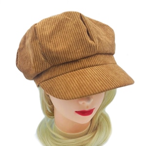 Può includere: Un berretto da giornalaio in velluto a coste marrone chiaro con una corona arrotondata e una visiera corta e rigida. Il cappello è esposto su una testa di manichino con capelli biondi e rossetto rosso. Il berretto ha creste verticali in velluto a coste.