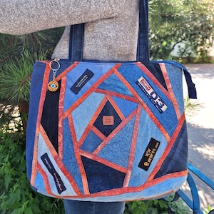 May include: A patchwork denim tote bag with a geometric design. The bag features various shades of blue denim, accented with orange stitching. Patches of fabric with text are visible. A small charm hangs from the handle.
