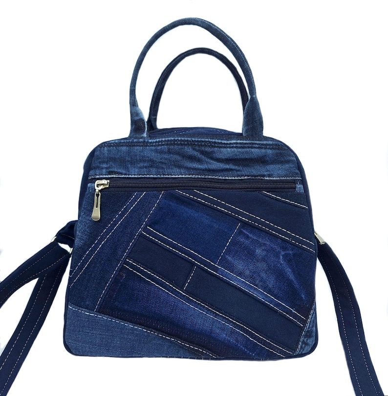 May include: A blue denim handbag featuring a patchwork design with various shades of blue fabric. The bag includes two top handles, a long shoulder strap, and a horizontal zippered pocket on the front with a metallic zipper pull.