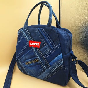 May include: A dark blue denim handbag featuring a patchwork design of various denim shades and textures. The bag includes two top handles, a shoulder strap, a small zippered front pocket, and a red Levi's brand patch on the front exterior.