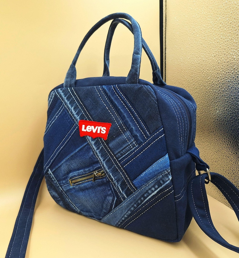May include: A dark blue denim handbag featuring a patchwork design of various denim shades and textures. The bag includes two top handles, a shoulder strap, a small zippered front pocket, and a red Levi's brand patch on the front exterior.