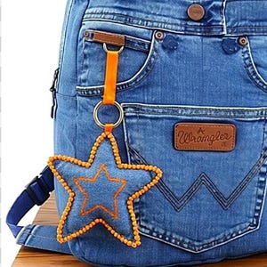 Può includere: Uno zaino in denim blu con una tasca frontale, una toppa in pelle marrone Wrangler e un portachiavi a forma di stella. Il portachiavi è realizzato in denim blu con cuciture arancioni e bordi di perline arancioni, fissato da un nastro arancione.